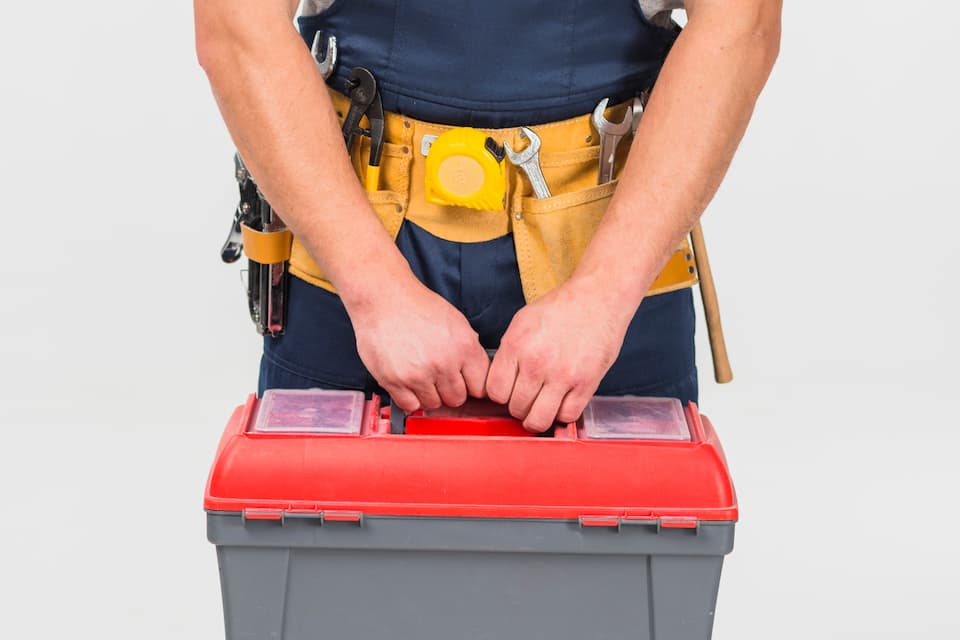 Hausmeisterservice: Eine Person im blauen Overall mit einem Werkzeuggürtel um die Hüften und eine rot - graue Werkzeugbox trägt.